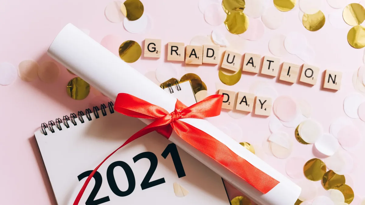 A white spiral-bound calendar with the year "2021" printed on it, a rolled-up piece of paper tied with a red ribbon, and the words "GRADUATION DAY" spelled out in letter tiles.